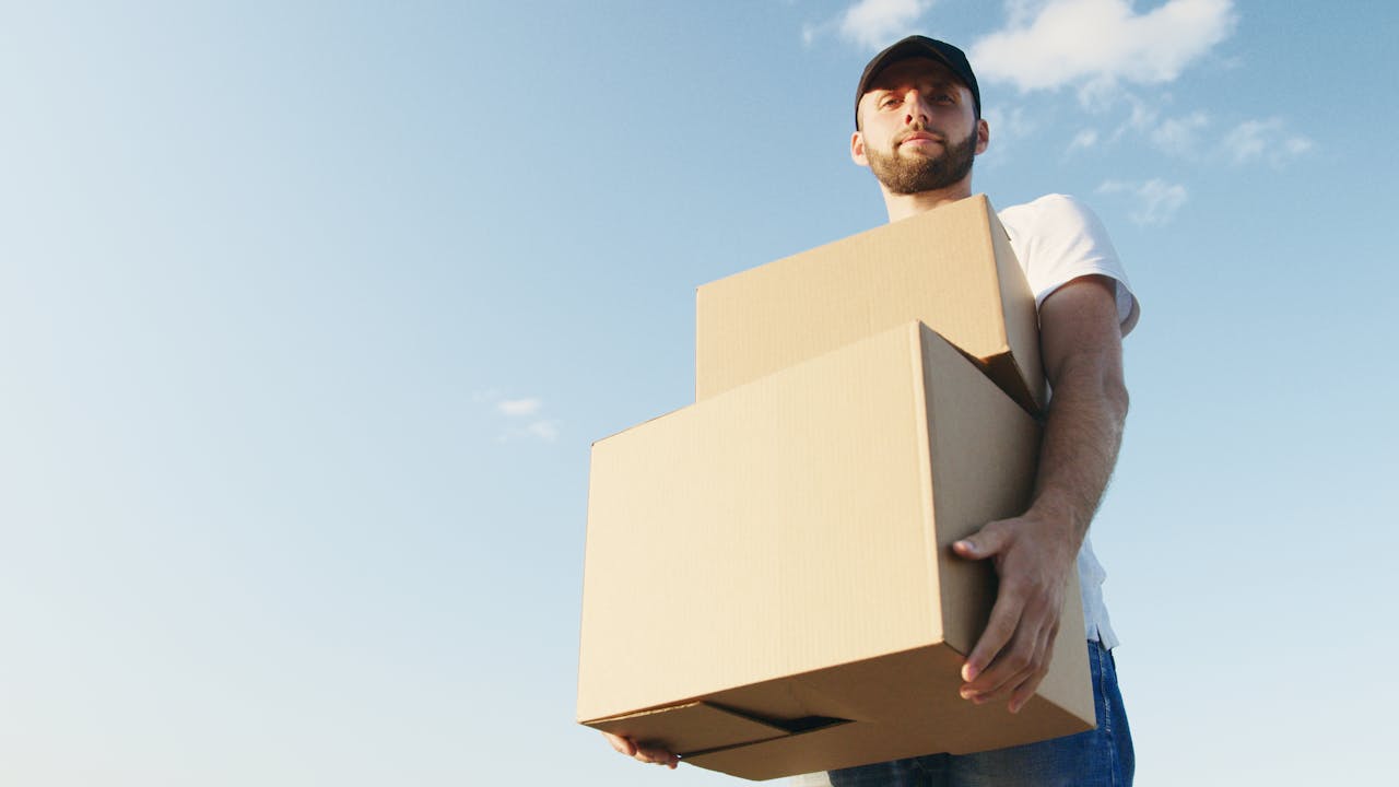 Man in casual attire with beard holding cardboard boxes under a bright blue sky, symbolizing delivery service.