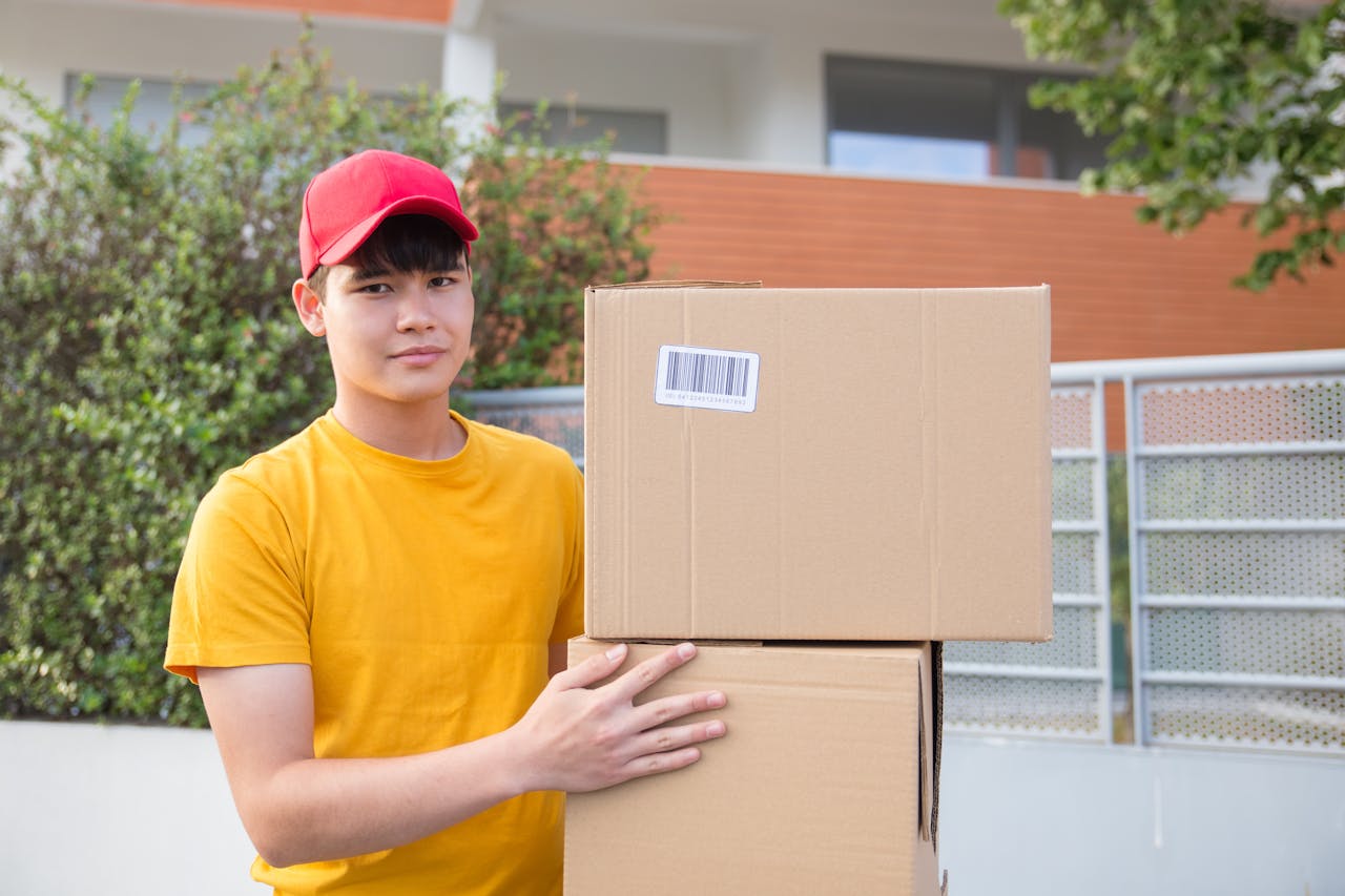 Asian delivery man holding cardboard boxes in an outdoor setting, wearing a red cap and yellow shirt.